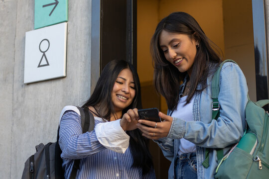 Young female university students sharing information on a mobile phone, happily connecting and using digital technology for education and social media networking outside, carrying backpacks