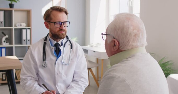 Senior man shares health concerns with male doctor during visit at hospital, GP professional asking about symptom or conditions, writing notes, filling medical form. Gerontology, healthcare services