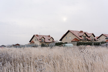 Suburban houses with red tile roofs behind frost-covered dry grass field on misty winter morning with pale sun in overcast sky. Concept of residential neighborhood, rural living and cold season.