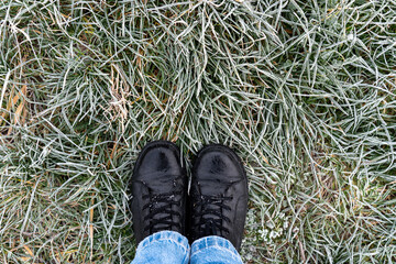 Top view of black leather boots and blue jeans standing on frost-covered green grass on cold winter morning. Concept of winter walk, outdoor activity, cold weather fashion and seasonal footwear.