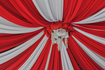 A visually striking pattern of alternating red and white draped fabric radiates from a central white floral chandelier, forming a canopy ceiling, symbolizing national colors, festivity, and wedding or