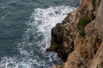 waves crash against rough, eroded limestone cliffs, creating white foam and deep blue water texture. a dramatic, high-angle close-up view of the adriatic sea