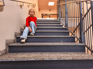 Young girl with blonde hair wearing a red sweater and blue jeans sits on stairs, resting her chin on her hand, reflecting a moment of contemplation in a modern indoor environment