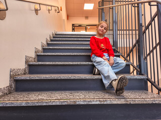 Young girl with blonde hair wearing a red sweater and jeans is sitting on stairs, enjoying a moment of relaxation in a modern indoor environment with a warm atmosphere
