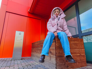 Young woman wearing a pink puffer jacket and blue jeans sits on a wooden bench against a vibrant red wall, showcasing modern urban fashion and style in a contemporary setting