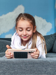 Young girl with long hair, wearing a white hoodie, is happily playing a game on her smartphone while lying on a cozy couch, showcasing joy and engagement in a playful environment