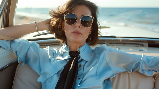 Fototapeta elegant woman with bobbed hair and sunglasses, wearing a light blue blouse and white trousers, sitting in the back seat of a vintage car near the sea