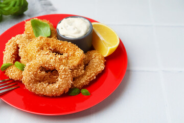 Deep fried squid rings with basil, sauce and slice of lemon on white tiled table, closeup. Space for text