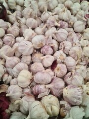 A close-up view of numerous garlic bulbs stacked together. The garlic has a pale purple skin with some white and brown tones visible.