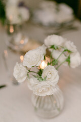 White roses in a small glass vase on an elegantly set dining table with warm candlelight