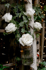 White roses in a glass jar hanging from a wooden structure surrounded by greenery and twinkling lights