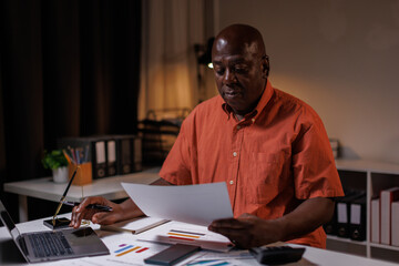 Senior businessman working late at night in his office, Senior man sitting at desk and talking on mobile phone while checking graphs on laptop.