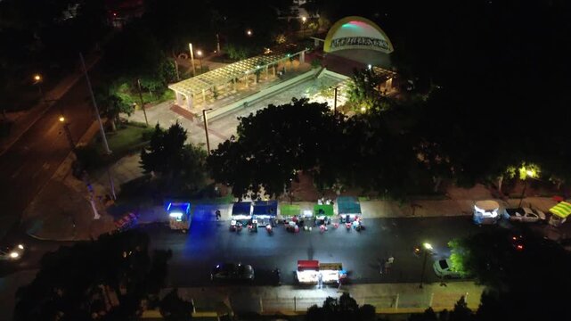 Parallel night aerial shot of street vendors beside Las Am&eacute;ricas park in M&eacute;rida, with festive lighting, food stands, and active nighttime movement. Captured with a 4K drone.