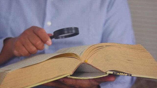 Searching for specific information or words in a book with a magnifying glass. A man opens a large book and uses a magnifying glass to clarify the information.Cognitive knowledge and educational learn