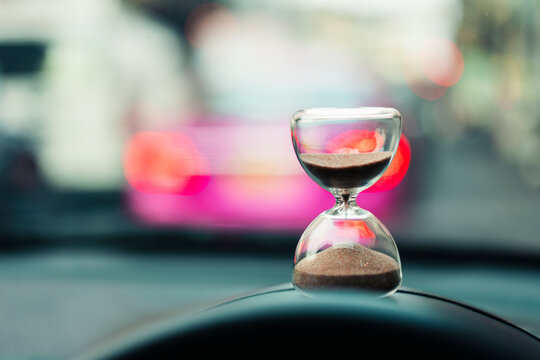 Hourglass placed on car dashboard with blurred traffic background, symbolizing time, waiting, patience, and delays during travel or commuting in modern urban lifestyle and busy city life.