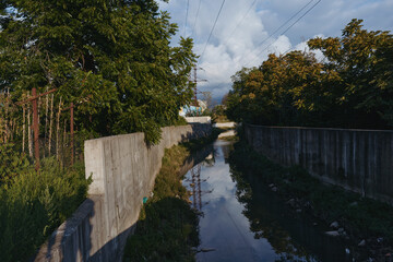 Fototapeta premium Concrete canal and urban drainage with narrow stream, overgrown vegetation and power lines reflected in water under moody sky at late afternoon, quiet industrial outskirts scene.