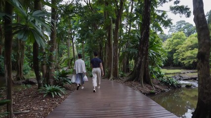 Two tourists are enjoying a leisurely walk along a wooden path, surrounded by the lush greenery of a tropical forest, creating a serene and peaceful atmosphere