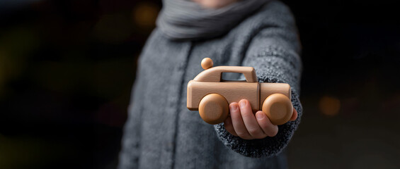 young child wearing gray sweater, confidently holding out a classic wooden toy car directly toward the camera, emphasizing early childhood learning
