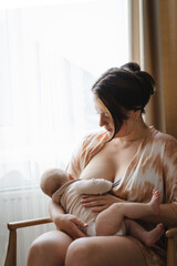 A mother sits in a wooden chair, gently breastfeeding her baby in a warm and softly lit room. Sunlight filters through the curtains, creating a peaceful atmosphere.
