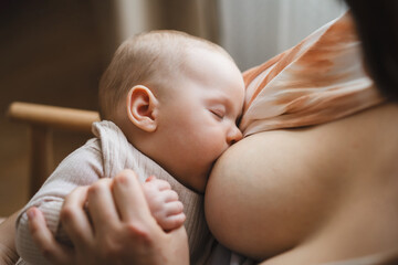 A mother sits in a wooden chair, gently breastfeeding her baby in a warm and softly lit room. Sunlight filters through the curtains, creating a peaceful atmosphere.