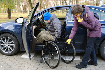 A woman holds a wheelchair with her right hand, a male driver transfers into a wheelchair from his car