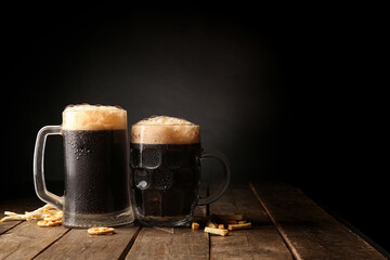 Mugs of dark beer and snacks on table against black background