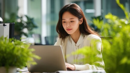 A young woman sits at a desk in a bright office space focused on her laptop. Green plants surround her creating a refreshing work environment during the day.