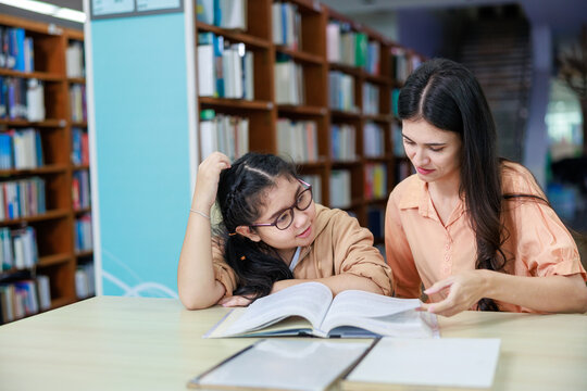 A young Asian girl reads a book with her teacher in a bright classroom, highlighting learning, guidance, and education in a warm, engaging school environment.