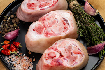 Three raw pork hocks displayed on a black plate with herbs, spices, and seasonings ready for...