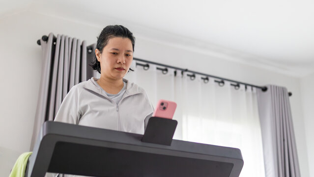 Focused Asian woman wearing wireless earbuds exercising on a treadmill indoors, using her smartphone for entertainment or tracking during her workout