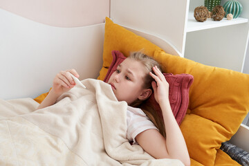 Sick teenage girl laying in bed and looking at a thermometer