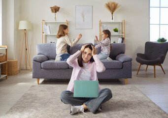 Sad frustrated mother trying to work on laptop, tired woman holding head with hands while two siblings play and have noisy pillow fight on sofa. Chaos and mess from kids activity in home living room