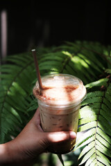 Close up hand holding a plastic glass of ice latte coffee with chocolate powder on topping in natural sunlight and tropical fern forest garden