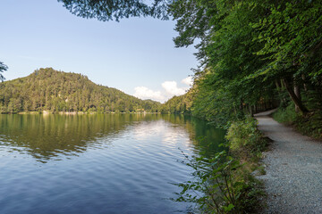 Tranquil Eibsee lake reflects forested hills under a summer sky, with a path winding along the shore inviting leisurely strolls