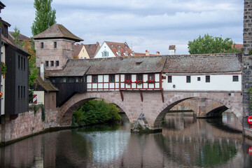 Historic stone bridge with wooden structure spans over tranquil river, surrounded by lush greenery and charming buildings, showcasing architectural beauty and serene landscape
