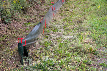 Green erosion control fabric secured with red stakes lies on the ground, surrounded by grass and wildflowers, showcasing environmental protection measures in landscaping