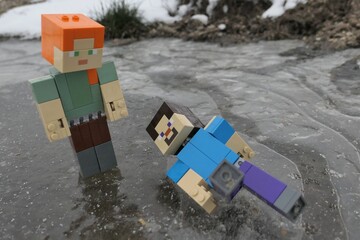 Naklejka premium LEGO Minecraft action figure of Steve laying on icy surface after fall while gliding with Alex, standing above him. Air bubbles visible under frozen ice puddle. 