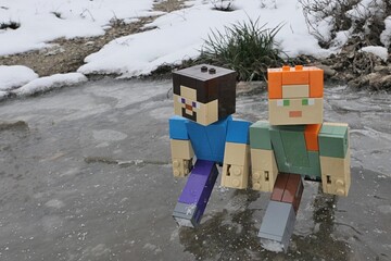 Naklejka premium LEGO Minecraft figures of main characters Steve and Alex gliding on ice of frozen puddle on edge of agricultural field, snowy puddle bank and frozen mud in background. 