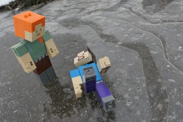 Naklejka premium LEGO Minecraft action figure of Steve accidentaly fallen on icy surface while gliding with Alex, standing above him. Air bubbles visible under frozen ice puddle. 