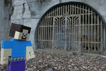 Naklejka premium LEGO Minecraft large figure of main character Steve standing in front of rusty gate to a mine or mining storage in village Hodrusa - Hamre, central Slovakia, opposite abandoned Sandrik factory