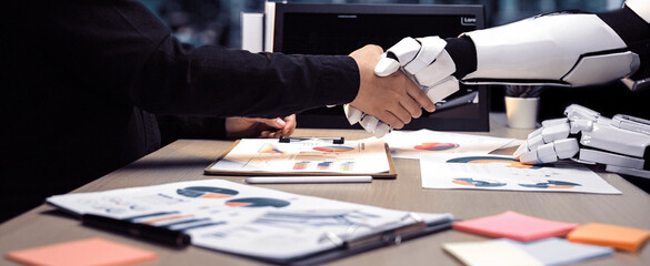 A human hand shakes with a robotic hand, representing a partnership in a modern office. Visual elements like charts and papers highlight the themes of innovation and collaboration.Noogenesis