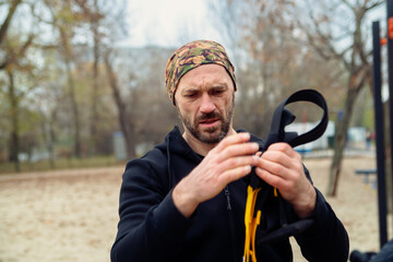 A focused man in a bandana and hoodie adjusts his suspension trainer straps outdoors, ready for a workout. Ideal for themes of fitness, outdoor exercise, and active lifestyles.