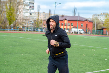 A focused bearded man in a black hoodie runs on a green stadium track during an outdoor workout, embodying an active, healthy lifestyle in an urban setting.