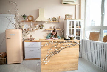 Young girl studying at a bright, cozy kitchen island adorned with spring cherry blossoms & Easter decor. A charming, light-filled domestic scene, perfect for themes of home, family, or springtime.