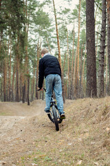 Fototapeta premium Young boy riding a BMX bike uphill on a dirt path through a serene pine forest, viewed from behind. Captures an active, adventurous outdoor moment amidst nature's tranquility.