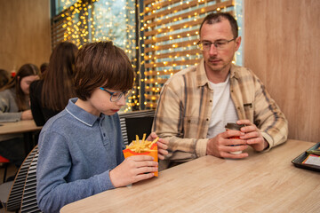 A father and son are sitting at a cafe table enjoying French fries and takeaway coffee, surrounded by warm string lights and a wooden interior.