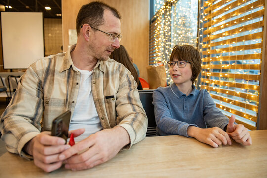A father and son are sitting at a table in a cozy cafe, illuminated by warm garlands in the background, and chatting casually, holding a smartphone in their hands.