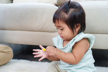 Fototapeta premium Adorable asian toddler girl sitting on floor holding colorful button in hand examining object curiously during early learning playtime at home representing exploration growth fine motor development