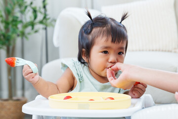 Adorable asian toddler girl sitting in feeding chair holding fork with strawberry being fed by parent learning interaction trust sensory development during early growth stage expressing bonding inside