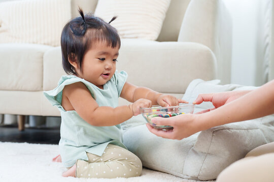 Adorable asian toddler girl sitting on floor playing with colorful buttons handed by adult during fun home learning activity expressing curiosity and joyful early childhood development moment